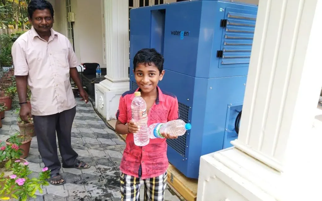A boy holding two bottles of water beside a Watergen unit in Kerala, India